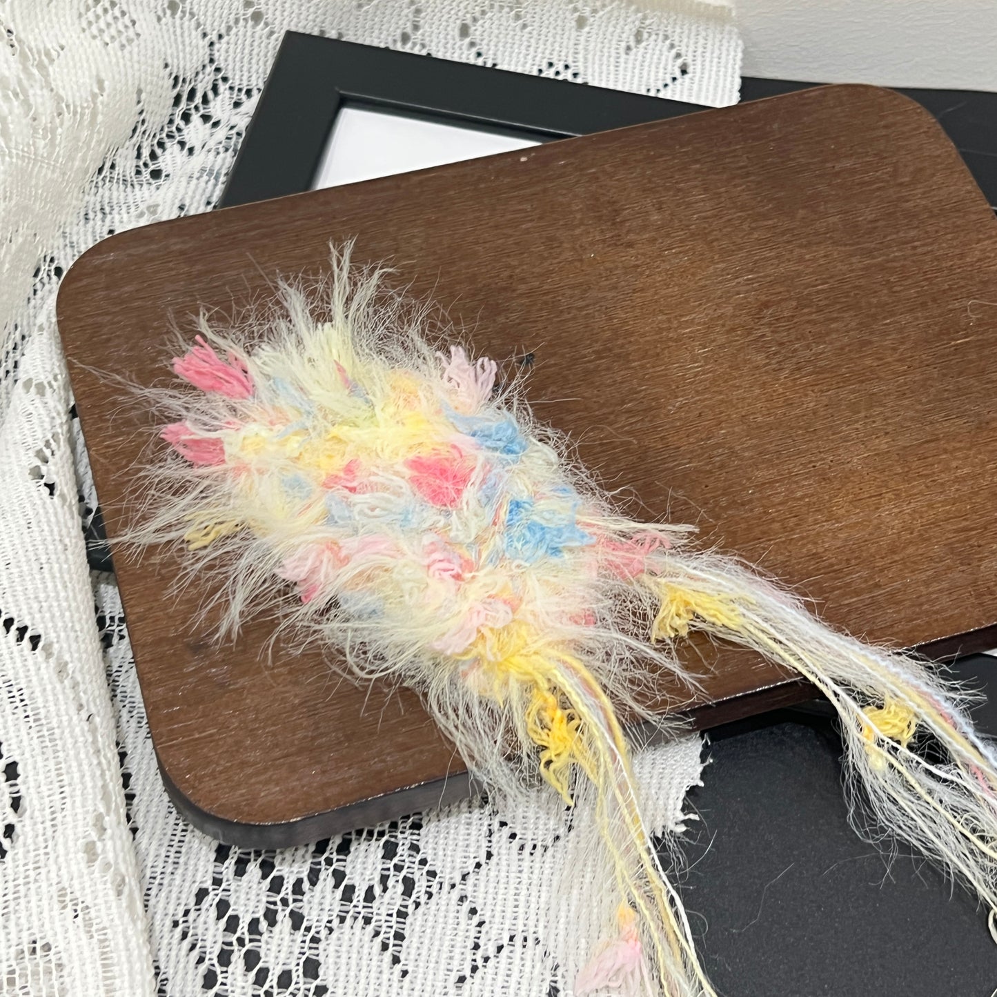 Close-up macro shot of cream pink blue fiber swatch laid flat on wooden board, long fuzzy feather yarn and multi-tone petal ribbon details clearly visible