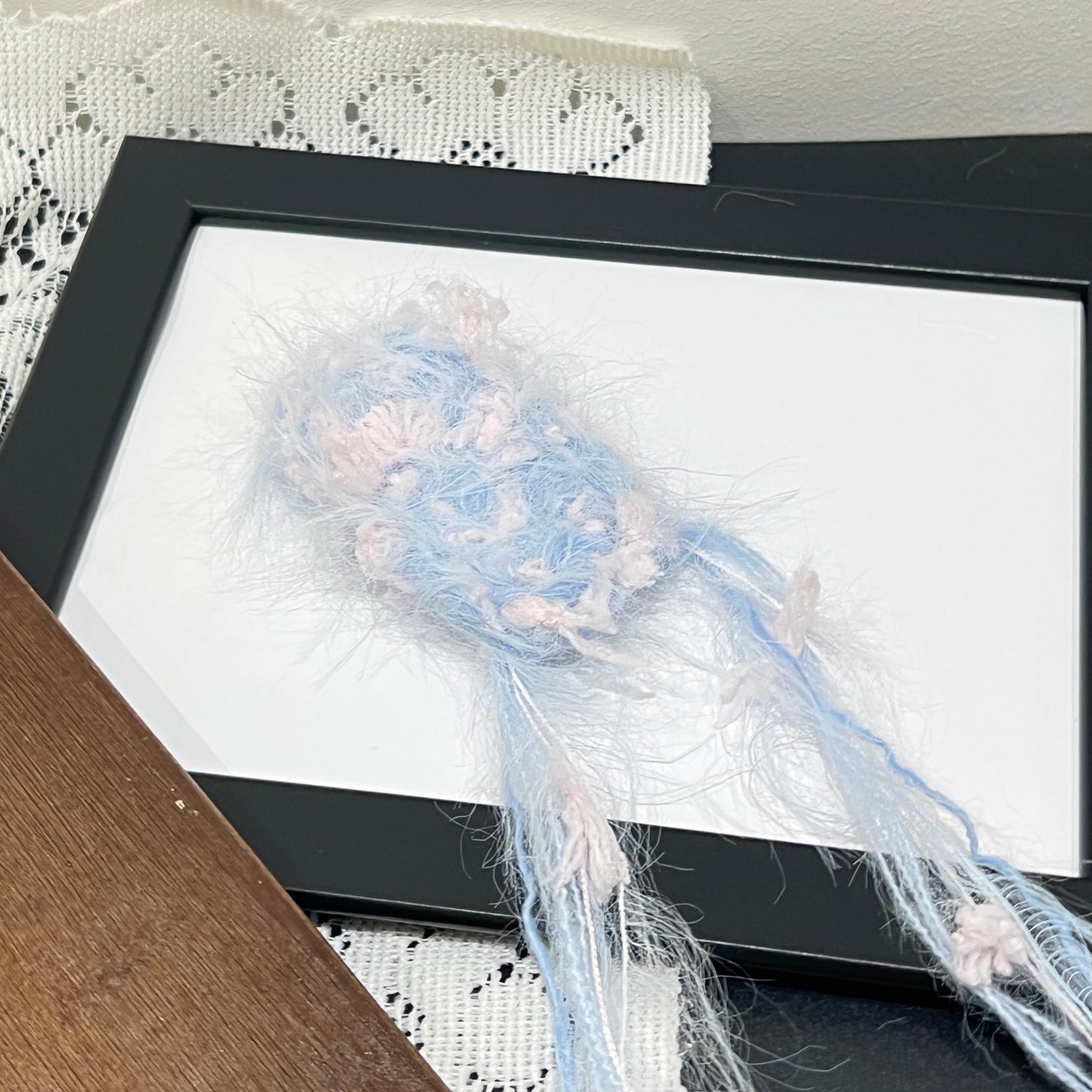 Close-up macro shot sky pink fiber swatch mounted inside thin black photo frame, long airy feather yarn and blush pink petal ribbon details