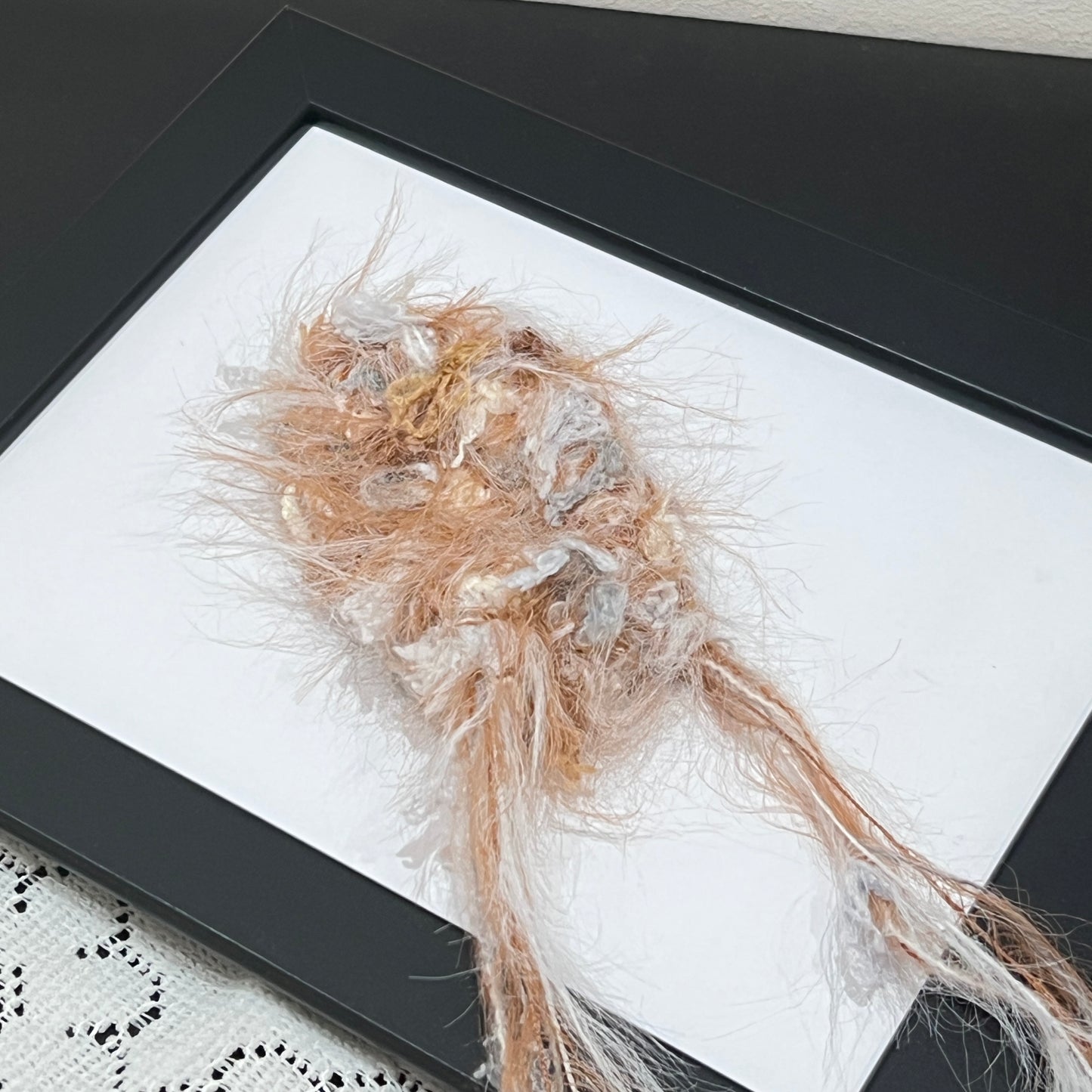Close-up macro shot caramel ivory fiber swatch mounted inside thin black photo frame, long fuzzy feather yarn and neutral tone petal ribbon details