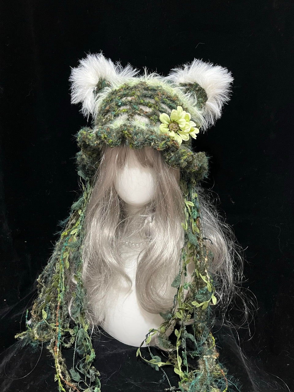 handmade crochet fairycore cat ear hat in moss green textured yarn, decorated with a flower and leafy vine ties, a woodland cosplay hat with fluffy white inner ears