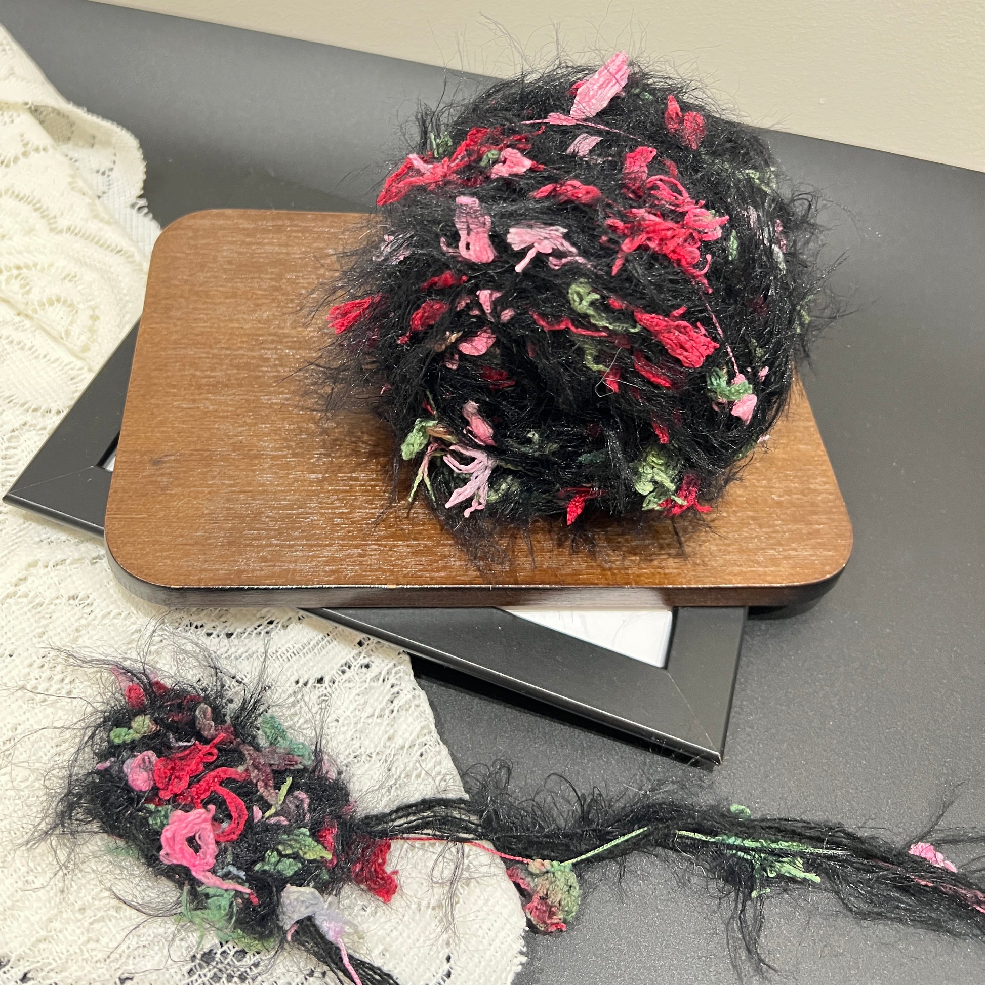 Full layout display set: black floral yarn cake + detached fiber swatch, perforated white lace fabric laid flat on dark matte countertop surface