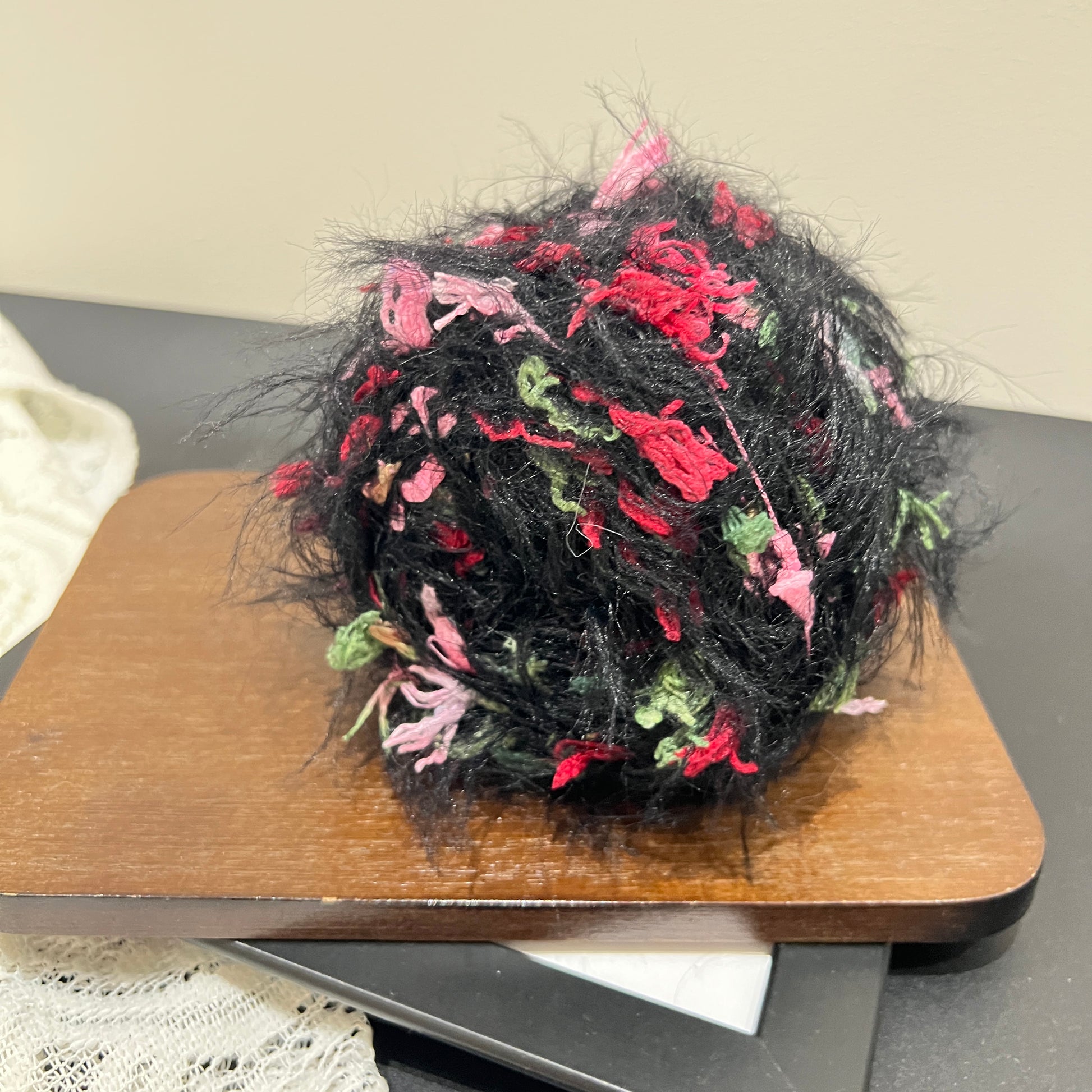 Side angle shot black blended yarn ball resting on square dark wooden display board, multi-color floral petal fluff clearly visible all over exterior