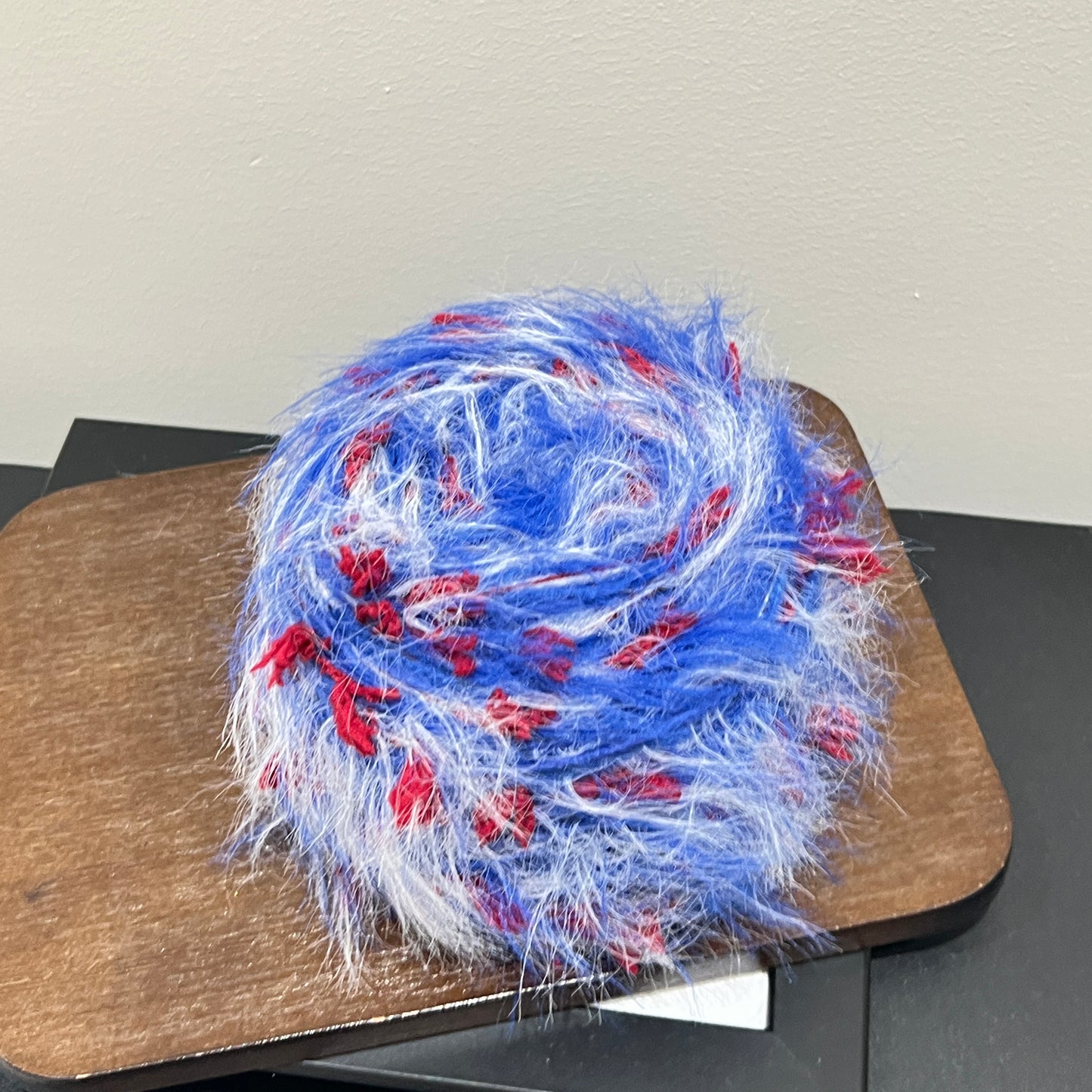 Side angle shot bright blue base blended yarn ball resting on square dark wooden board, tri-color petal fluff clearly visible around exterior