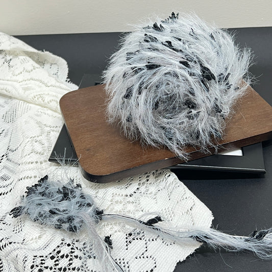 Front overhead spherical shot pale gray multi-strand hand-blended yarn cake, scattered solid black petal ribbon fiber accents, airy long layered feather texture, wooden display board, white lace fabric & neutral light wall backdrop