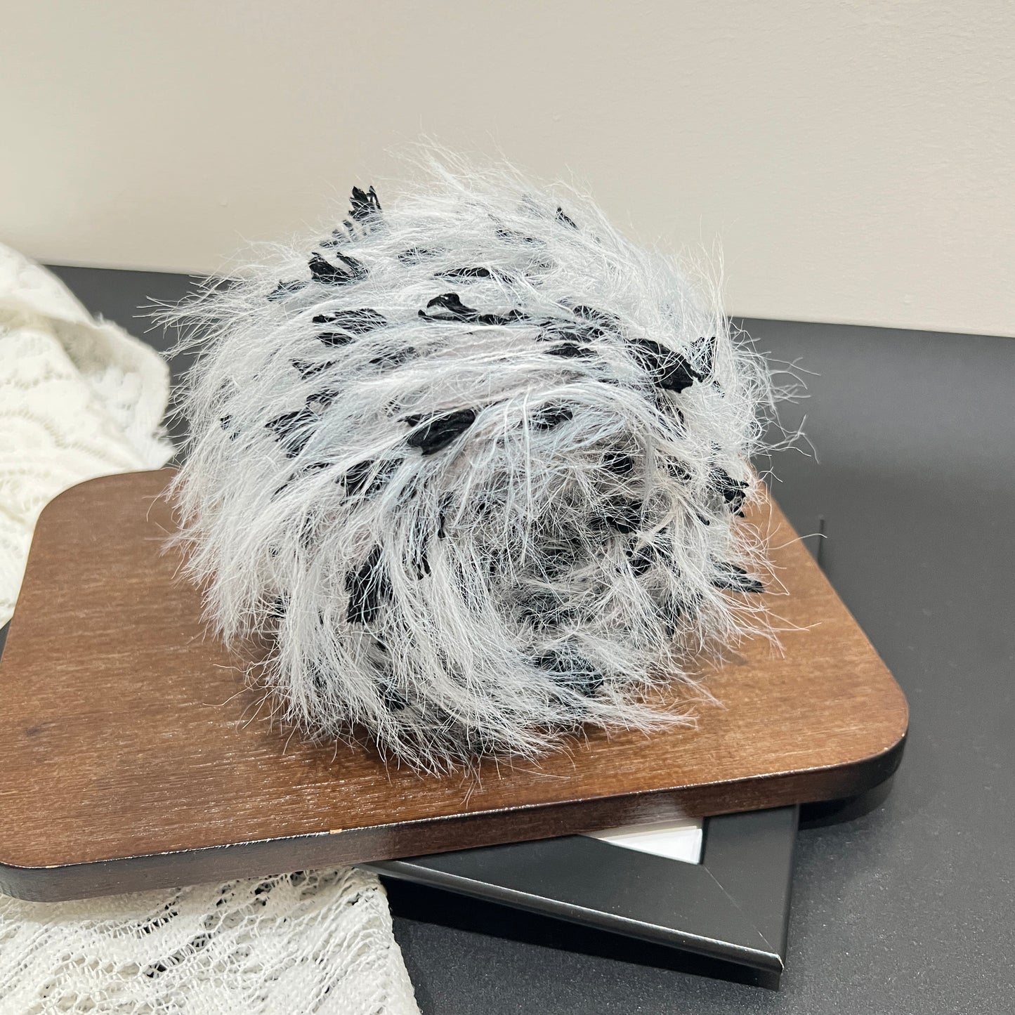 Side angle shot pale gray blended yarn ball resting on square dark wooden board, distinct black petal fiber details all around exterior