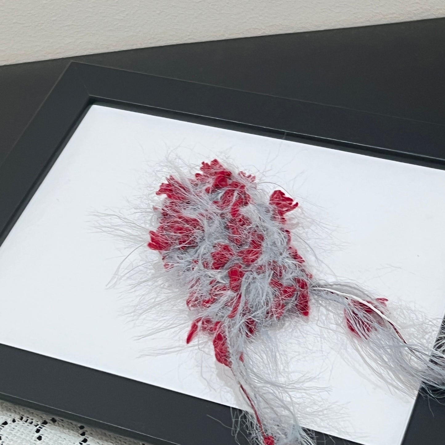 Close-up macro shot gray scarlet fiber swatch mounted inside thin black photo frame, long airy feather yarn and vivid scarlet petal ribbon decorative details