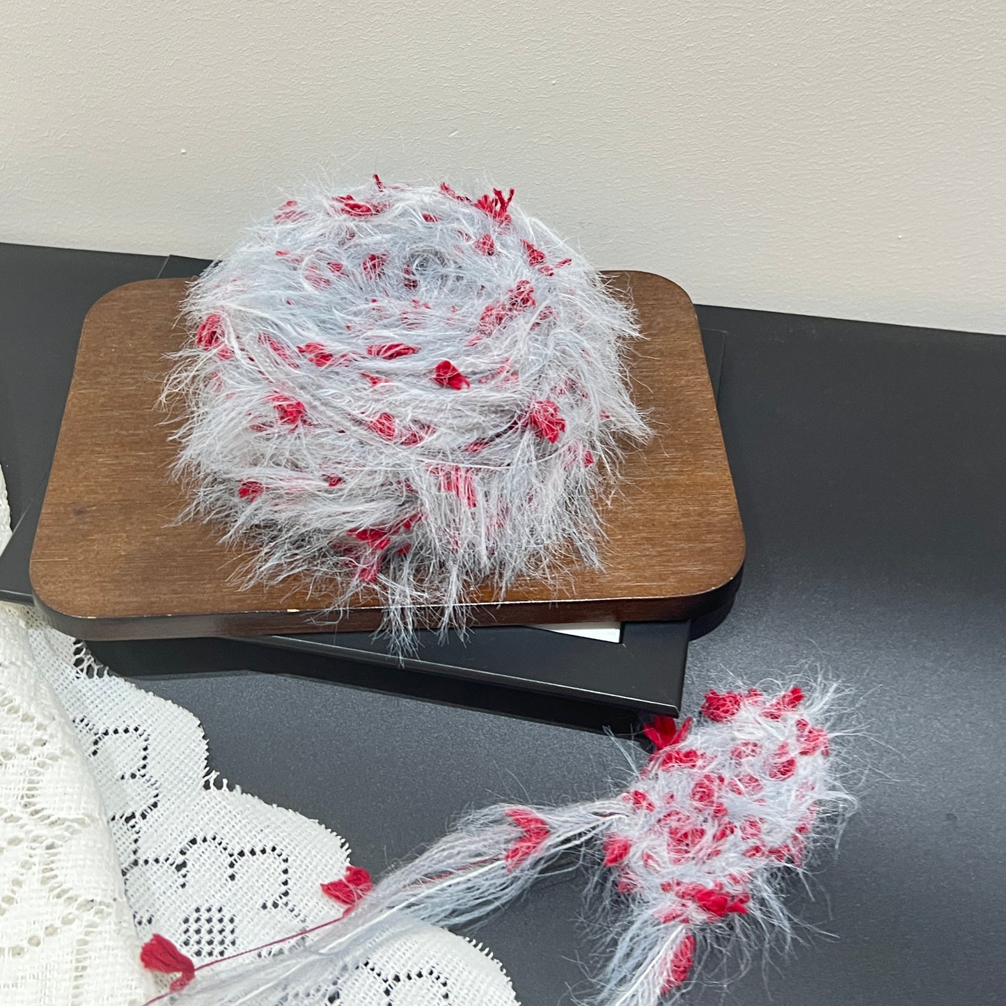 Front overhead spherical shot pale gray floral multi-strand hand-blended yarn cake, scattered scarlet red petal fiber accents, airy layered feather texture, placed on wooden board with plain neutral wall backdrop