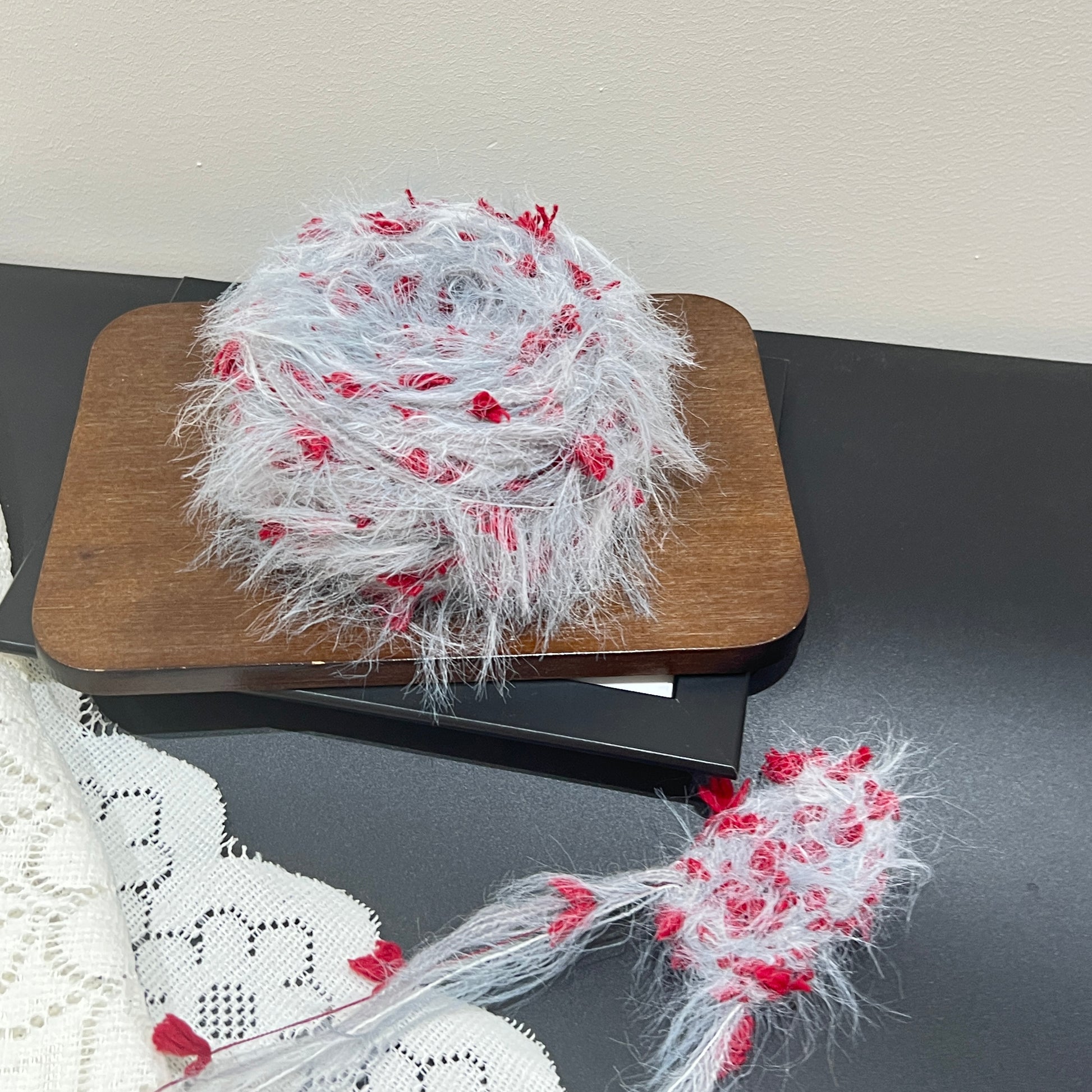 Front overhead spherical shot pale gray floral multi-strand hand-blended yarn cake, scattered scarlet red petal fiber accents, airy layered feather texture, placed on wooden board with plain neutral wall backdrop