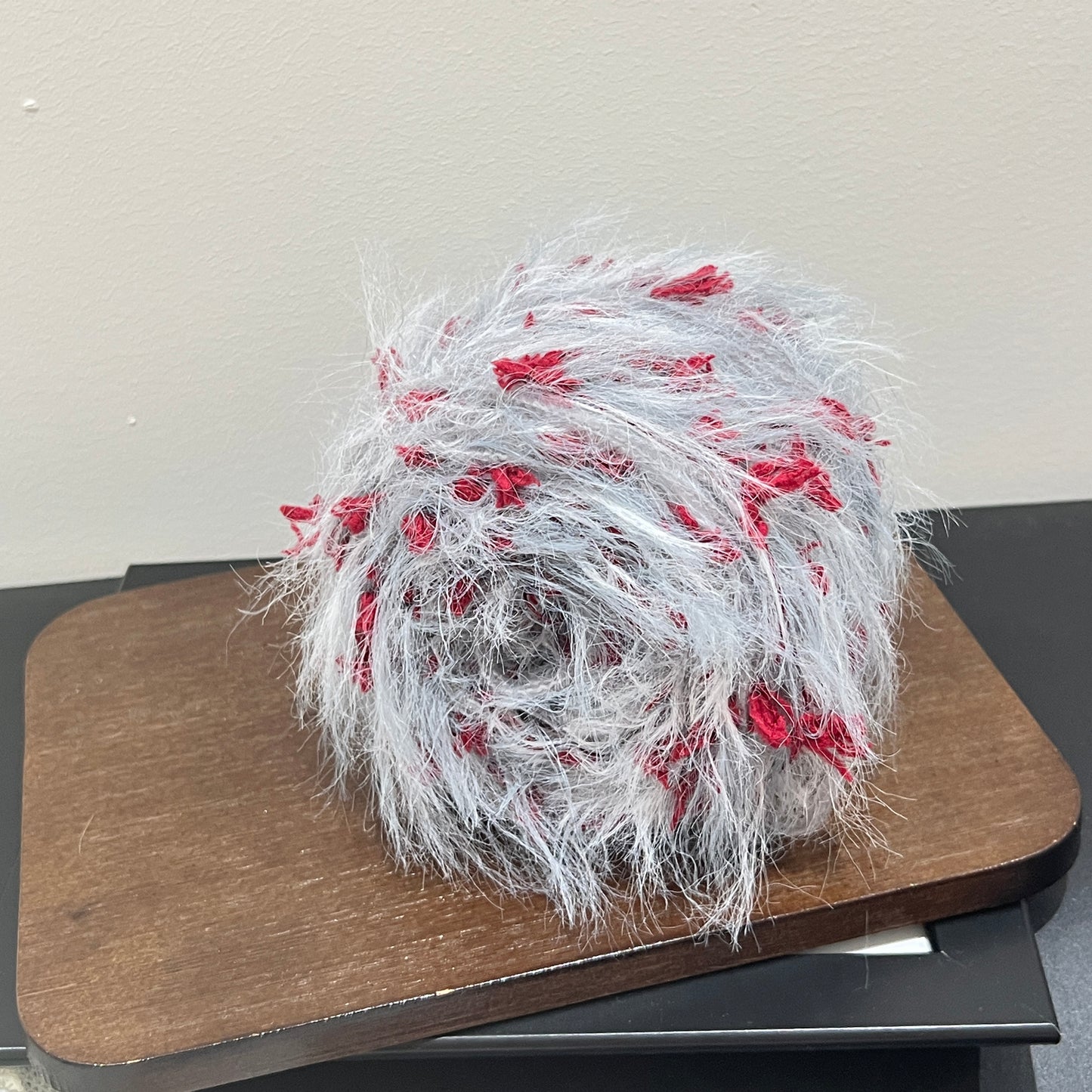 Side angle shot pale gray blended yarn ball resting on square dark wooden display board, scarlet red petal fluff clearly visible all over surface