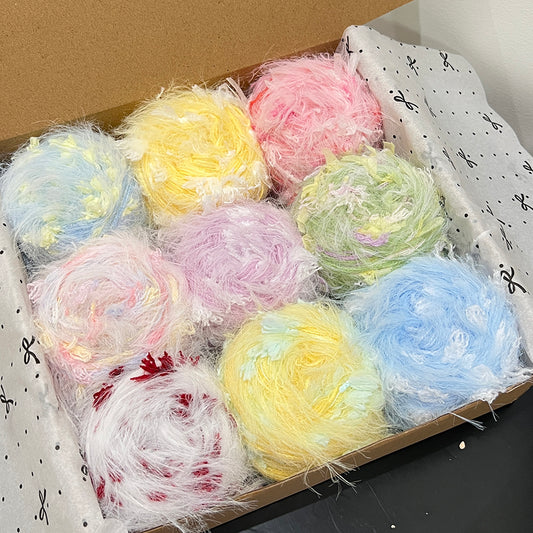 Open gift box containing 9 assorted pastel floral hand-blended yarn cakes, soft sky blue, blush pink, lavender, yellow, mint green, white crimson color palette, wooden gift box with printed tissue paper, dark table backdrop