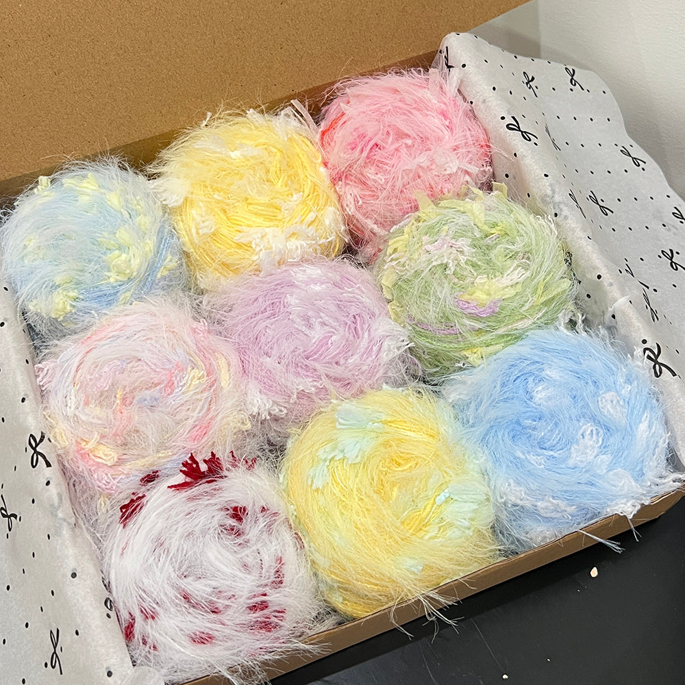 Open gift box containing 9 assorted pastel floral hand-blended yarn cakes, soft sky blue, blush pink, lavender, yellow, mint green, white crimson color palette, wooden gift box with printed tissue paper, dark table backdrop