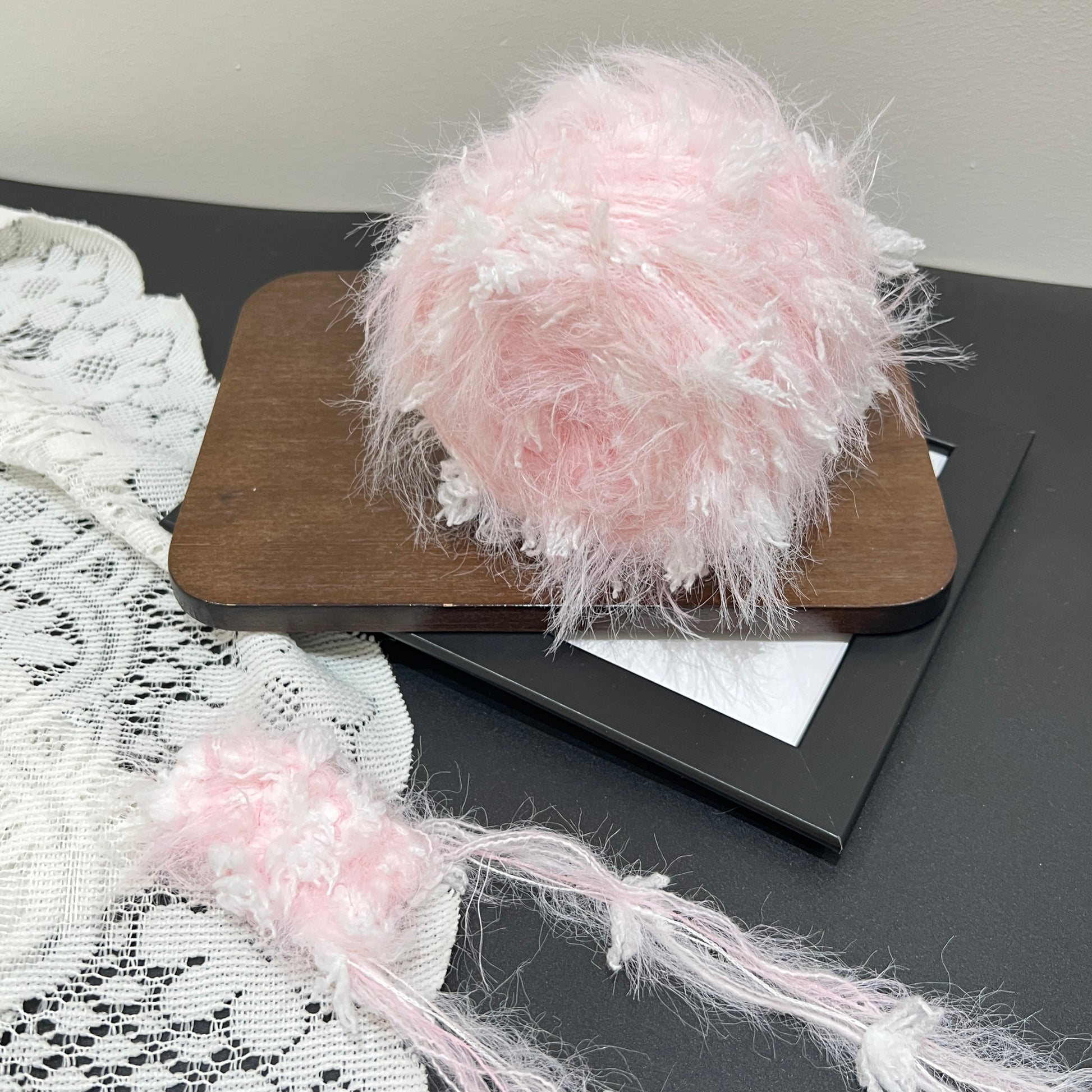 Full display of pastel blush yarn cake and matching fiber swatch on dark surface with lace decor