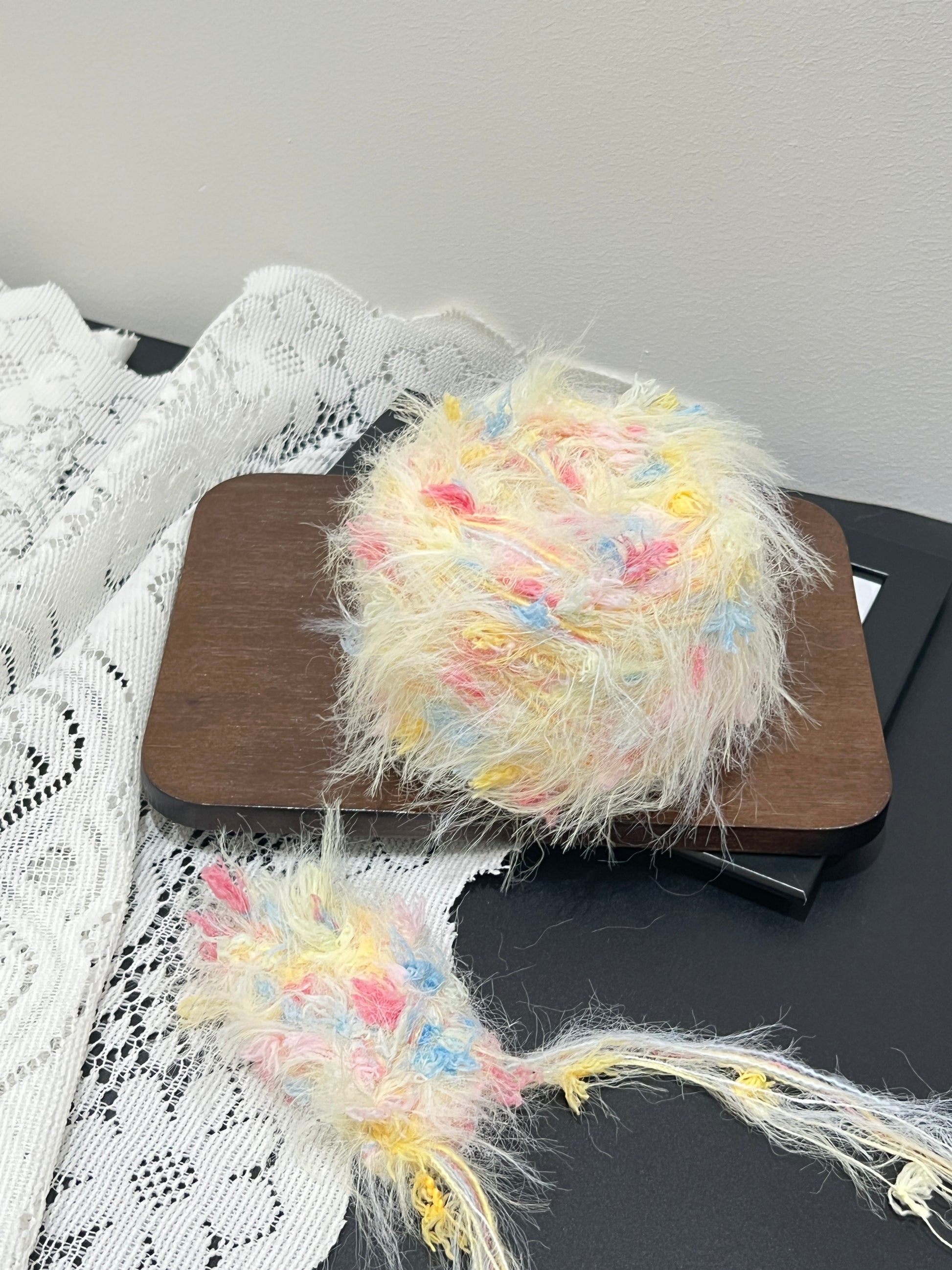 Full layout display: cream yellow yarn ball + detached fiber swatch, hollow white lace fabric on left, dark matte countertop surface, neutral wall background