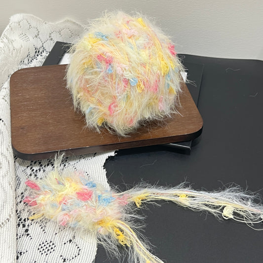 Front overhead spherical shot of pastel cream yellow floral multi-strand hand-blended yarn cake, decorated with scattered pink, sky blue petal fiber accents, airy layered feather texture, placed on wooden board with lace fabric backdrop