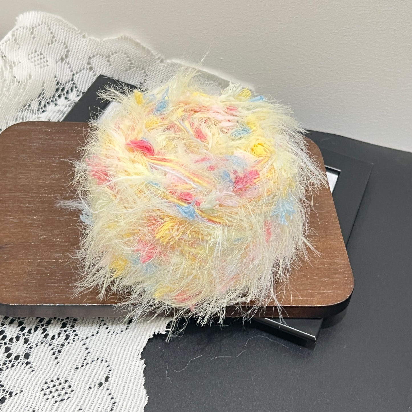 Side angle shot of cream base blended yarn ball resting on square wooden display board, multi-color pastel petal fiber details covering the surface