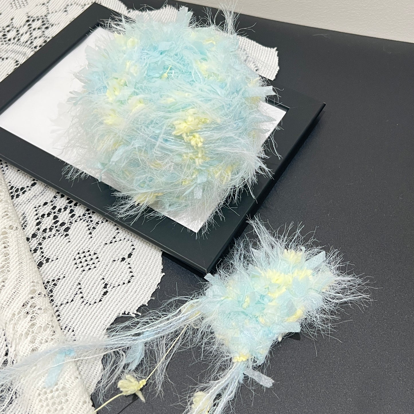 Pastel cyan yellow floral multi-strand hand-blended cake yarn placed on black frame with white lace backdrop