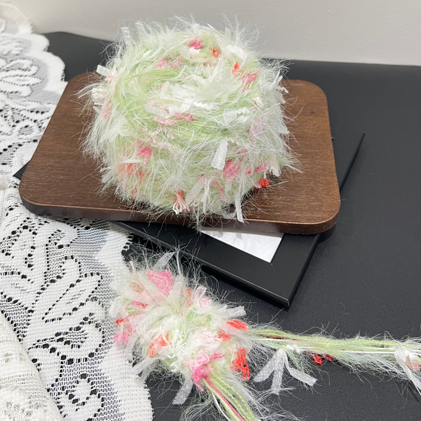 Full layout shot of pastel lime pink coral floral blended yarn cake and detached fiber swatch, laid on dark board with hollow white lace fabric, neutral wall backdrop