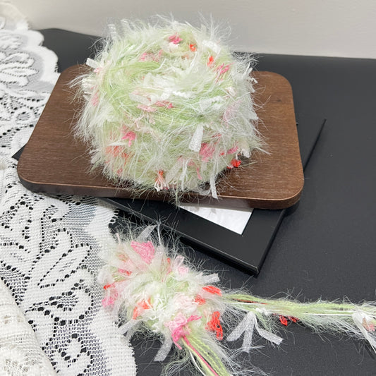 Full layout shot of pastel lime pink coral floral blended yarn cake and detached fiber swatch, laid on dark board with hollow white lace fabric, neutral wall backdrop