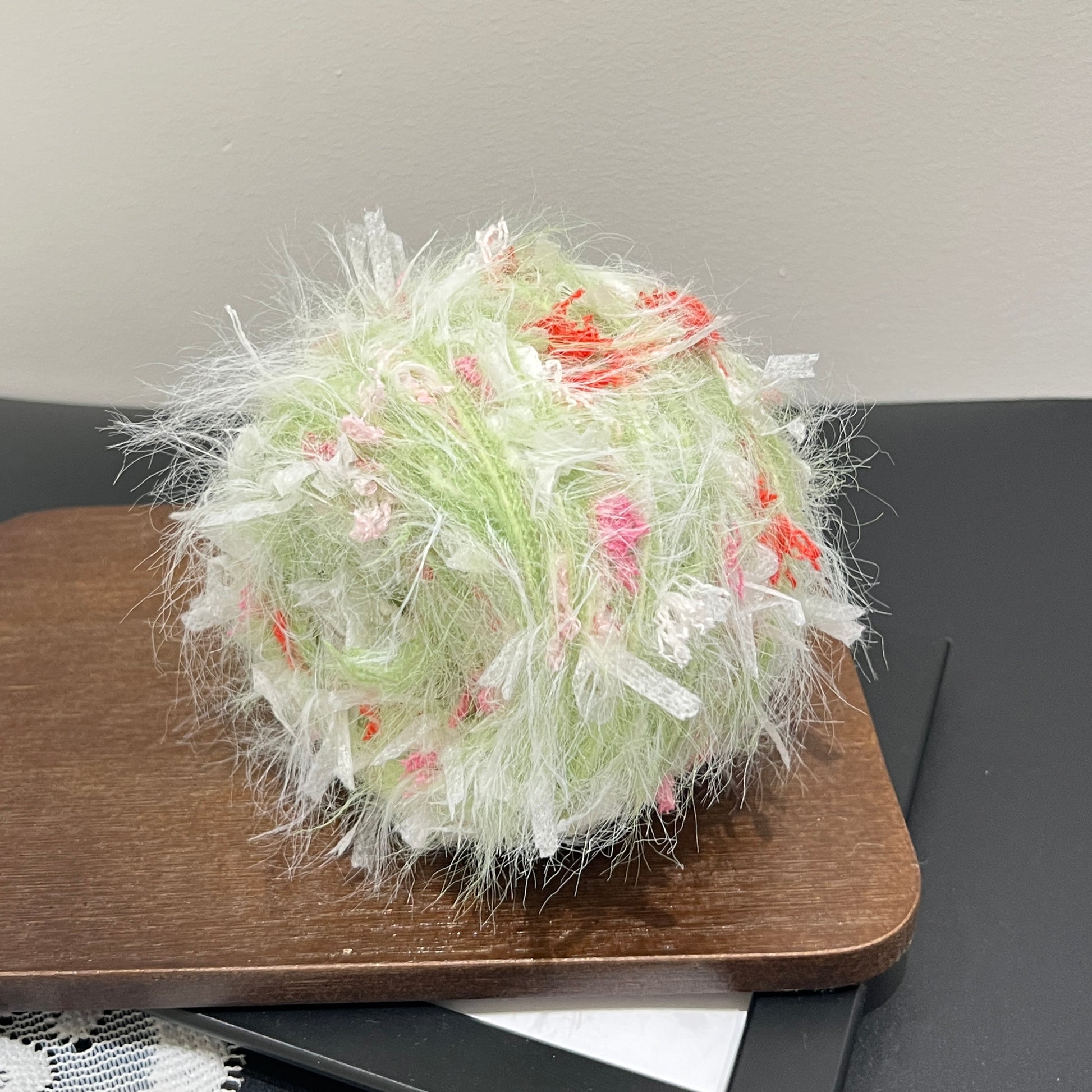 Side angle shot pastel lime yarn ball resting on wooden square board, multi-color petal fluff clearly visible around surface