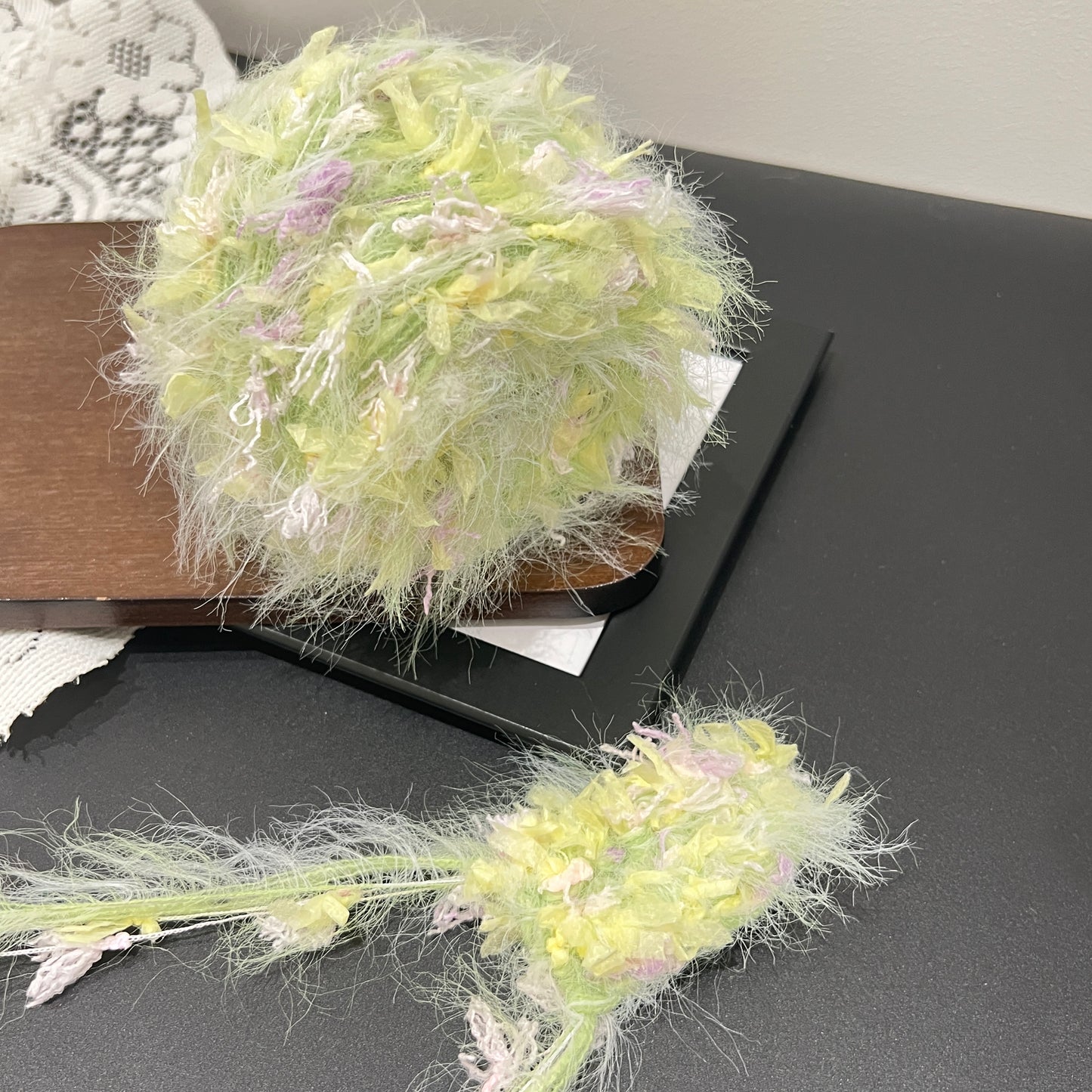 Side layout shot yarn ball + matching fiber swatch beside hollow white lace fabric on dark countertop