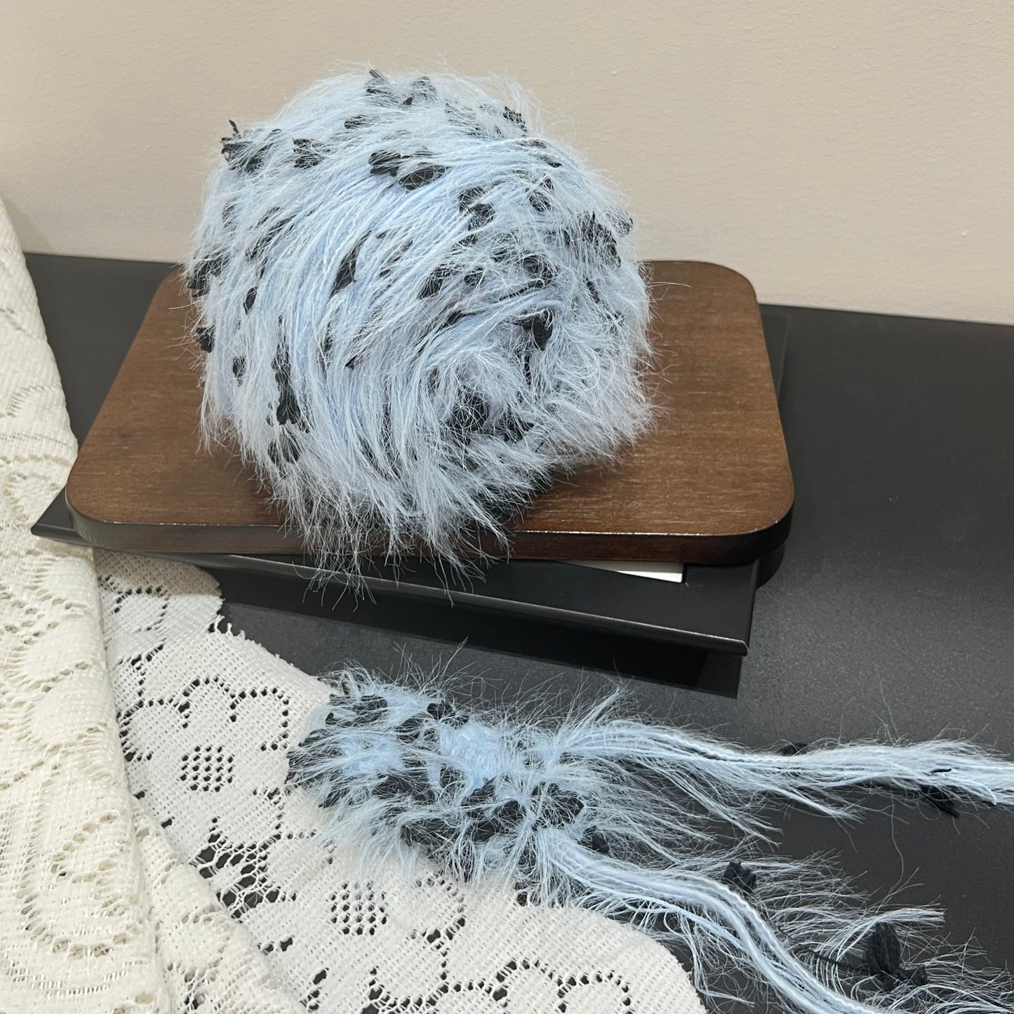 Front overhead spherical shot pastel sky blue multi-strand hand-blended yarn cake, scattered solid black petal ribbon fiber accents, airy long layered feather texture, wooden display board, neutral light wall background