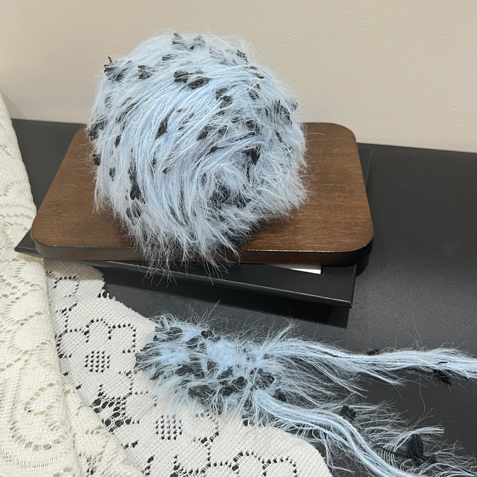 Front overhead spherical shot pastel sky blue multi-strand hand-blended yarn cake, scattered solid black petal ribbon fiber accents, airy long layered feather texture, wooden display board, neutral light wall background
