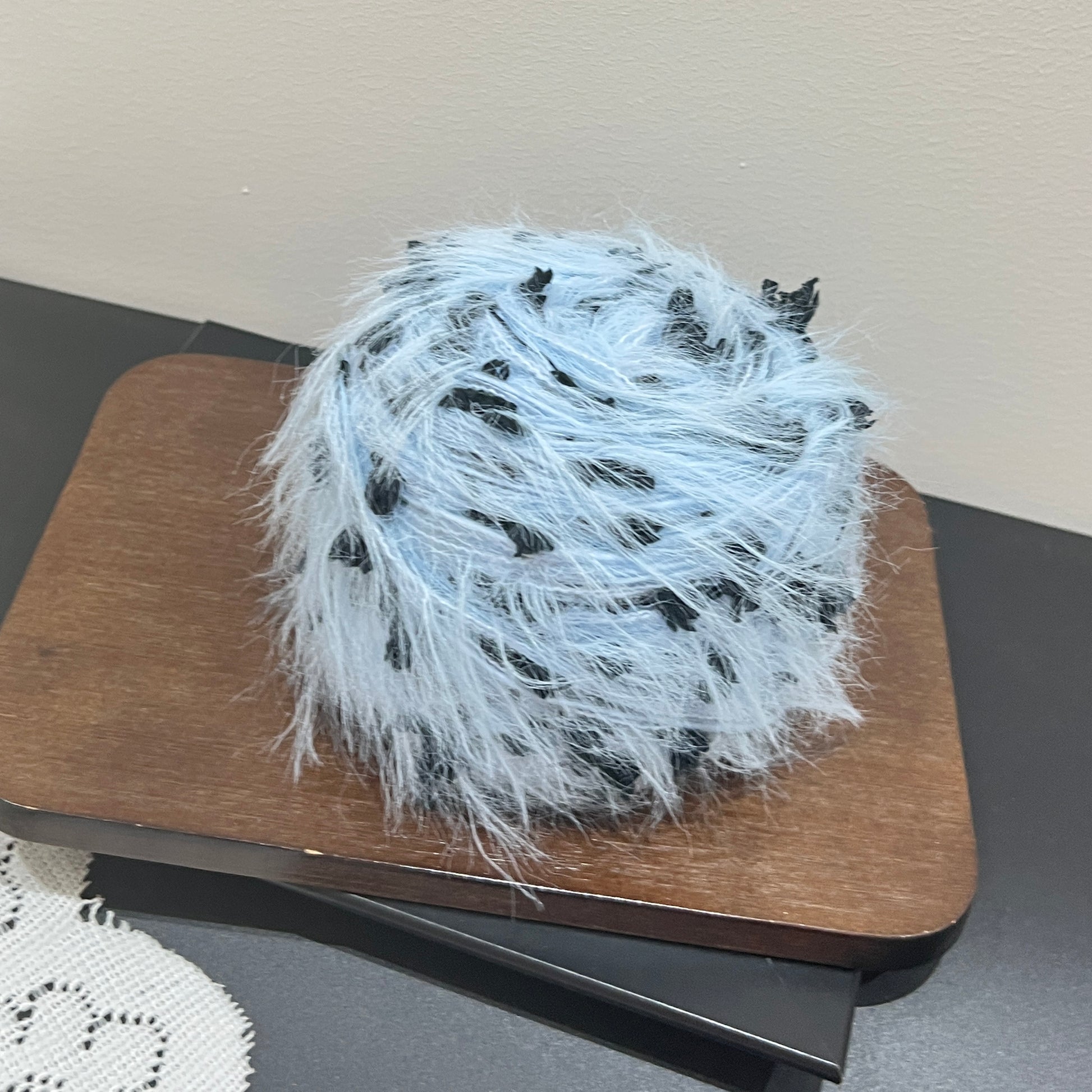Side angle shot pastel sky blue blended yarn ball resting on square dark wooden board, distinct black petal fiber details all around the exterior