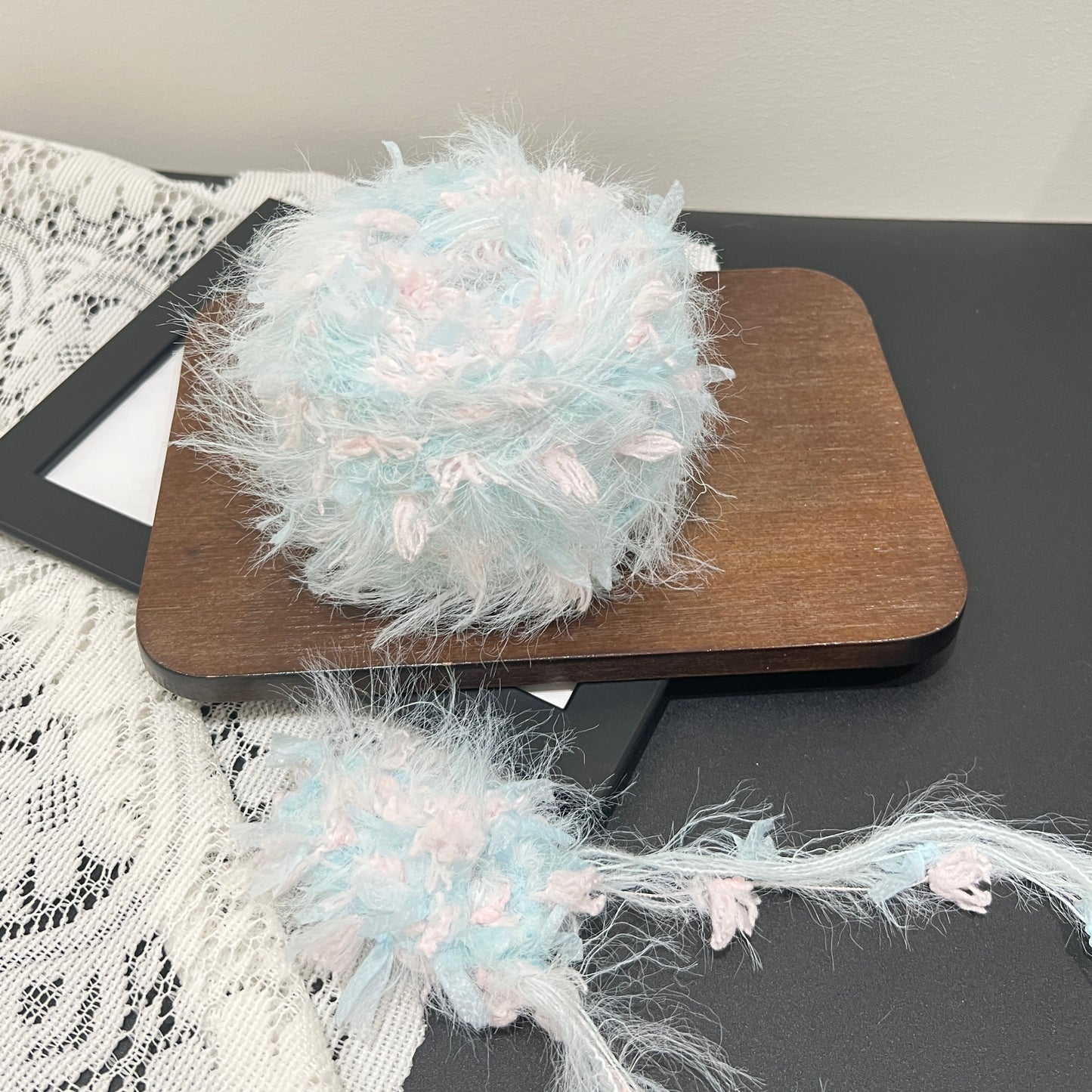 Pastel sky blue blush pink floral multi-strand hand-blended cake yarn ball placed on dark wooden square board, hollow white lace fabric on left side, plain neutral wall background