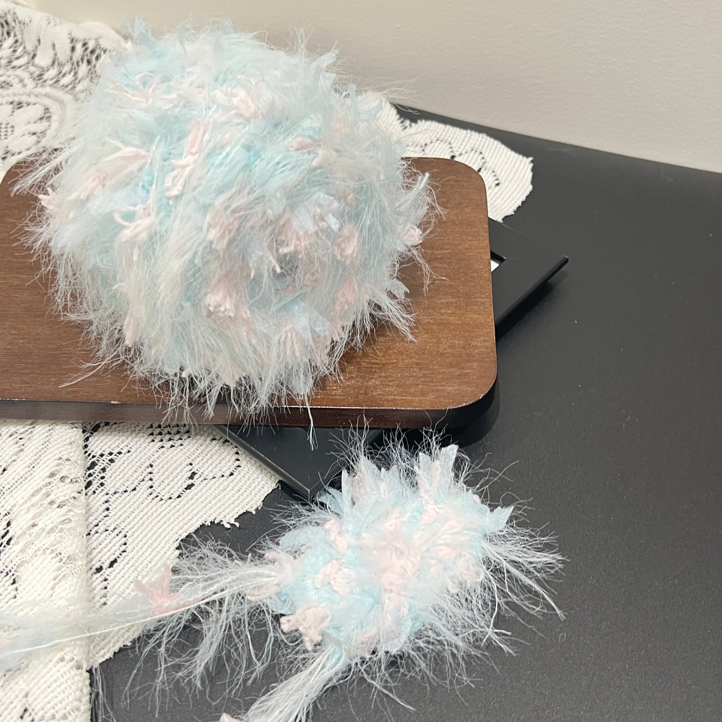 Close-up fiber swatch of pastel sky blue blush pink floral multi-strand blended yarn mounted on wooden display board, featuring long airy feather fiber and scattered pink petal ribbon accents