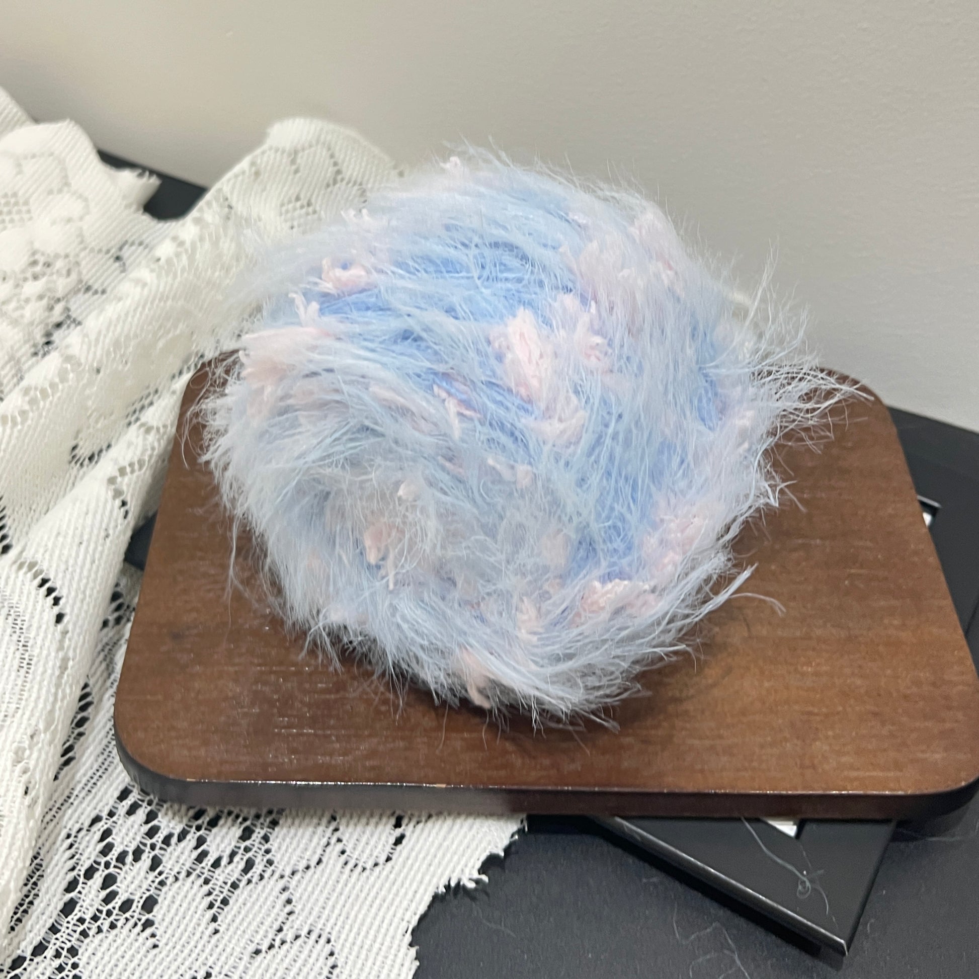 Side angle shot pastel sky blue yarn ball placed on square wooden board, multi-tone pink petal fluff distributed over surface