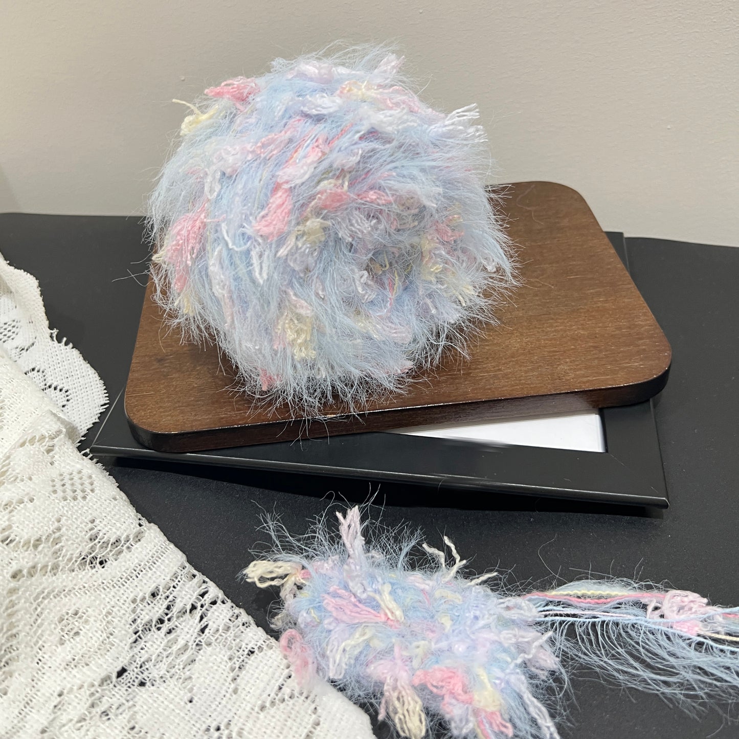 Pastel sky pink cream floral multi-strand hand-blended cake yarn placed on wooden board with white lace fabric backdrop