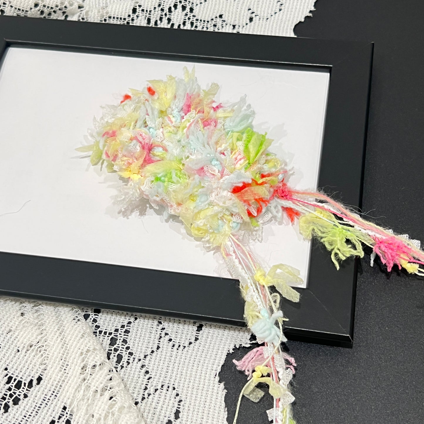 Close-up macro shot spring multi-pastel fiber swatch mounted inside thin black photo frame, layered shredded ribbon and soft textured decorative yarn details