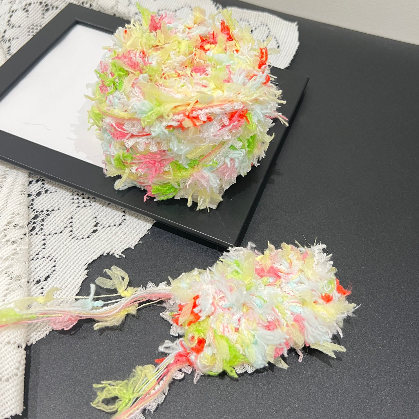 Full layout display set: spring multi-pastel yarn ball + detached long tassel fiber swatch, hollow white lace fabric laid flat on dark matte tabletop