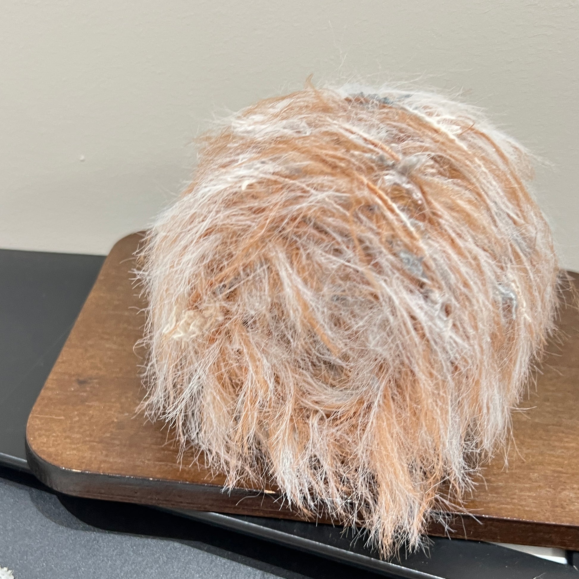 Side angle shot warm caramel base blended yarn ball resting on square dark wooden board, mixed neutral petal fluff distributed over entire surface