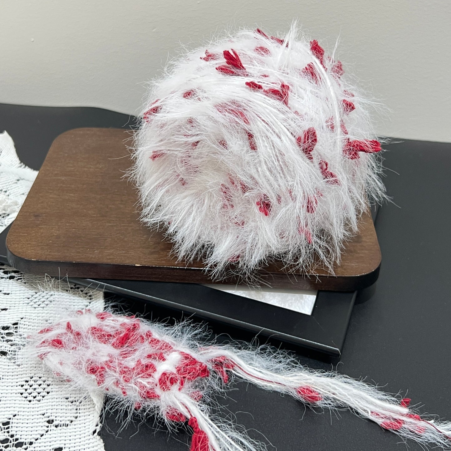 Layout shot of white crimson floral yarn cake and matching fiber swatch beside hollow white lace fabric on dark countertop